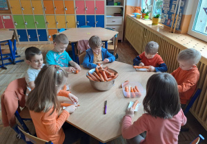 Dzieci siedzą przy stoliku i obierają marchewki, na środku stoi miska z obranymi i umytymi marchewkami.