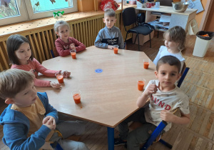 Dzieci siedzą przy stoliku i mają przed sobą sok marchwiowy w kubeczkach.
