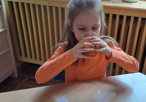 Dziewczynka siedzi przy stoliku i pije sok marchwiowy.