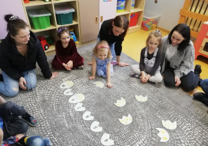 Rodzice i dzieci siedzą na dywanie, a jedna z dziewczynek wybiera połowę pisanki z kurczaczkami.