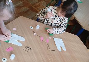 Dziewczynki wykonują papierowego zajączka. Przyklejają na wcześniej przygotowanym szablonie elementy przedstawiające zajączka.