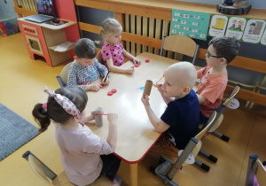 Piętka dzieci siedzi przy stoliku, cześć z nich smaruje klejem papierowe rurki po papierze toaletowym, a pozostali rysują kropki mazakiem na czerwonych nakrętkach – biedronki.