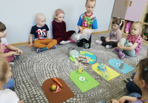 Dzieci siedzą w kole pośrodku, którego leży papierowa Ziemi przykryta odpadami oraz papierowe kosze w różnych kolorach, chłopiec trzyma w ręku folię i zastanawia się na który kosz ja położyć.