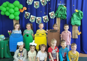 Dzieci z grupy Motylki pozują do zdjęcia. Czworo dzieci ma na szyi kolorowe kartony – oznaczające pojemniki do segregacji obok stoi narrator. Przed nimi siedzą dzieci dwoje dzieci ma opaski na głowie.