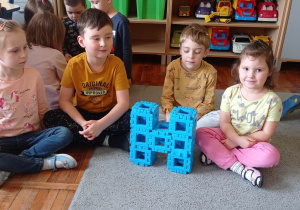 Czwórka dzieci siedzi na dywanie , pozuje do zdjęcia ze zbudowaną przez siebie literką „H” z klocków typu wafle.