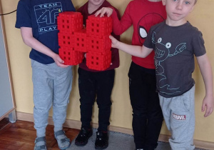 Czwórka dzieci pozuje do zdjęcia ze zbudowaną przez siebie literką „ H” z czerwonych klocków typu wafle.