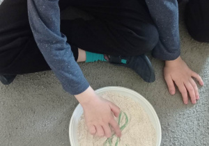 Chłopiec w niebieskiej bluzce prowadzi palcem po śladzie pisanej literki „H” zrobionej z plasteliny i ułożonej na talerzyku wypełnionym mąką.