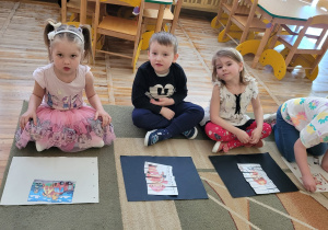 Dziewczynki i chłopiec siedzą na dywanie i prezentują ułożone obrazki