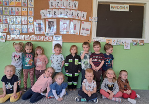 Dzieci z grupy Krasnale pozują do zdjęcia na tle napisu Dzień Strażaka. Jedna dziewczynka ubrana jest w strój strażaka.