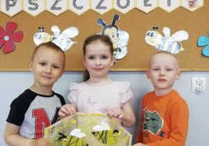 Trójka dzieci – dwóch chłopców i dziewczynka trzymają wykonaną przez siebie pracę plastyczną - plaster miodu z pszczółkami.