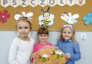 Trzy dziewczynki trzymają wykonaną przez siebie pracę plastyczną - plaster miodu z pszczółkami.