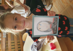 Dziewczynka prezentuje wykonany przez siebie portret swojej mamy.