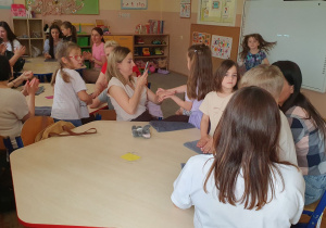 Zdjęcie przedstawia mamy siedzące przy stolikach oraz dzieci wykonujące im relaksacyjny masaż dłoni z wykorzystaniem kremu do rąk.