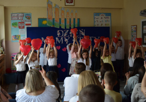Dzieci tańczą, trzymając nad głowami czerwone balony. Na widowni siedzą rodzice.