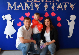 Rodzice wraz z dzieckiem na tle dekoracji pozują do rodzinnego zdjęcia. Rodzice w rękach trzymają upominki, a chłopiec dwa czerwone balony.