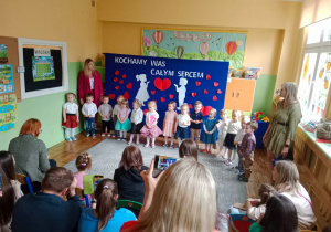 Szczęśliwe przedszkolaki recytują wierszyki dla rodziców w Sali pełnej gości na tle udekorowanego parawanu.