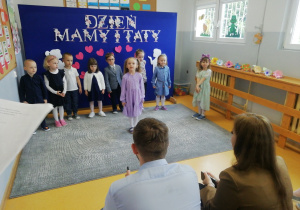 Dziewczynka w fioletowej piosence mówi wiersz dla rodziców.