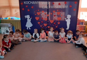 Dzieci z grupy stokrotki czekają na swoich rodziców siedząc na dywanie przed tablicą z dekoracją.