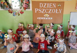 Dzieci z grupy Stokrotki pozują do zdjęcia na tle dekoracji z okazji Dnia Pszczoły, ze swoimi wcześniej zrobionymi pszczółkami.