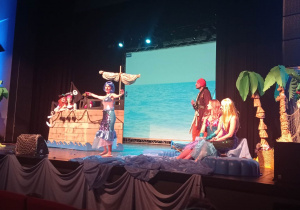 Na scenie stoi atrapa statku, obok stoi aktor odgrywający rolę pirata, po prawej stronie siedzą aktorki przebrane za syreny a jedna z nich wygłasza swoją rolę.
