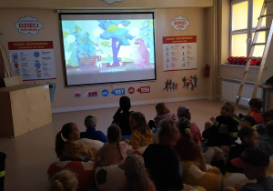 Dzieci siedzą na pufach w kształcie piłek i oglądają film edukacyjny.