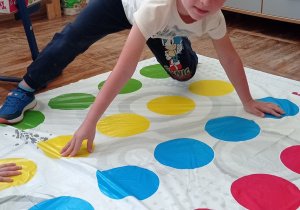 Zdjęcie przedstawia chłopca, który wykonuje zadanie ruchowe wyznaczone w grze pt.: „Twister”.