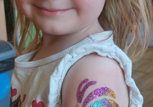 Uśmiechnięta dziewczynka pokazuje kolorowy tatuaż brokatowy, który ma na rączce.