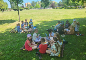 Dzieci z grupy Odkrywcy wybrały się do parku na piknik i jedzą smakołyki.