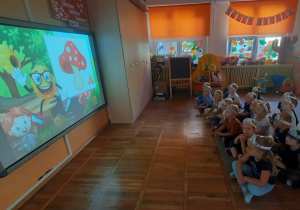 Dzieci z grupy Żabki siedzą na dywanie i oglądają na tablicy multimedialnej film edukacyjny o kropce.
