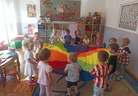 Dzieci stoją w kole trzymając w rękach kolorową chustę w kształcie kropki.