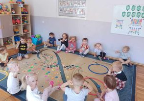 Dzieci siedzą na dywanie, na którym rozsypane są kolorowe kropki.