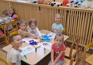 Dzieci wykonuja prace plastyczne siedząc przy stolikach.