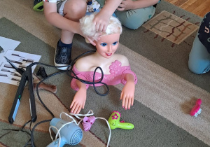Chłopiec upina włosy lalce, reszta dzieci siedzi w kole na dywanie.