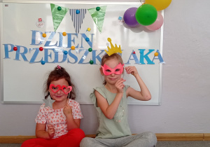 Dwie dziewczynki siedzą przed tablicą, pozują do zdjęcia z gadżetami: koroną, maskami na oczy, gwiazdką.