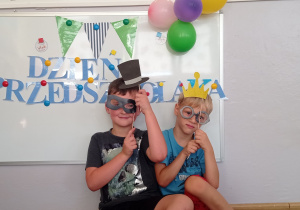 Dwóch chłopców siedzi przed tablicą, pozują do zdjęcia z gadżetami trzymając maski na oczy, koronę oraz czapeczkę.