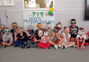 Grupa siedemnastu przedszkolaków siedzi przed tablicą pozując do zdjęcia z wykonanymi przez siebie maskami.
