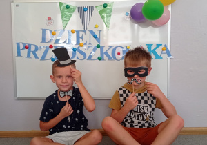 Dwóch chłopców siedzi pod tablicą pozując do zdjęcia z gadżetami: maską, muszką, okularami, wąsami.