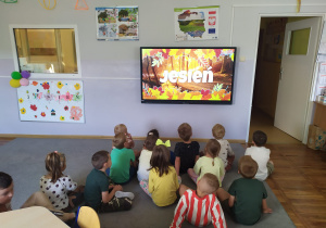 Dzieci siedzą przed projektorem multimedialnym i oglądają film edukacyjny pt. Kazio i jesień.