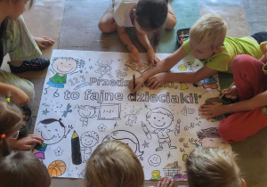 Dzieci wspólnie malują kredkami dużą malowankę „Przedszkolaki to fajne dzieciaki.”