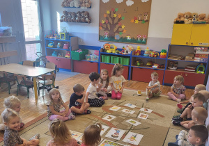 Dzieci siedzą na dywanie i oglądają obrazki przedstawiające charakterystyczne cechy jesieni.