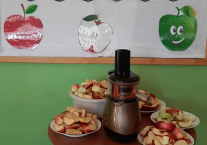 Tablica dekoracyjna z napisem Dzień Jabłka, przed tablicą stoi stolik na którym stoją talerzyki oraz miska z jabłkami i wyciskarką do soków.