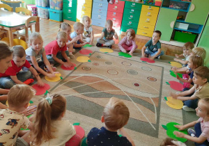 Dzieci wraz z Panią siedzą w kółku na dywanie, przed sobą mają jabłka z papieru i stukają w nie dwoma rękami.