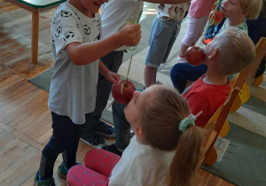 Z prawej strony dzieci siedzą na krzesełkach, po lewej stronie dzieci stoją i w rękach trzymają jabłka na sznurku.