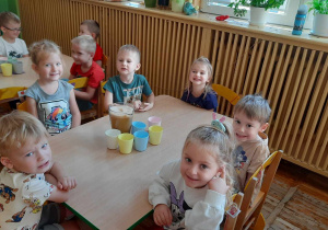 Dzieci siedzą przy stoliku na którym stoi dzbanek z sokiem oraz kubeczki.