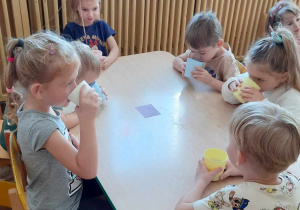 Dzieci siedzą przy stoliku i piją sok z jabłka w kubeczkach.