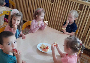Dzieci siedzą przy stoliku na którym stoi talerz z jabłkami oraz je zjadają.
