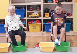 Dwójka chłopców bierze udział w zawodach w przenoszeniu piłeczek nogami z jednej miski do drugiej.