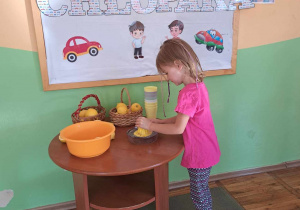 Na tle tablicy dekoracyjnej dziewczynka wyciska sok na wyciskarce, na stoliku stoją kubeczki, miska oraz koszyki z cytrynami.