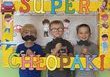 Na tle tablicy dekoracyjnej trzech chłopców pozuje do zdjęcia w fotobudce z napisem Super Chłopaki, w rękach trzymają papierowe wąsy.