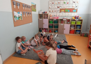 Dzieci leżą oraz siedzą na dywanie i słuchają bajki czytanej przez uczennicę klasy trzeciej.